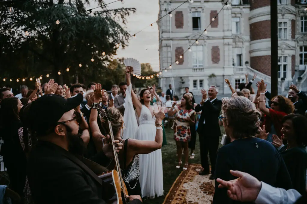 Voici une photo de mariage capturant la mariée dansant avec élégance, comme dans une danse espagnole, le bras levé et éventail à la main. Entourée de ses invités, elle profite d’un moment joyeux tandis qu’un guitariste-chanteur crée une ambiance festive devant le château du Parc Saint-Lambert.