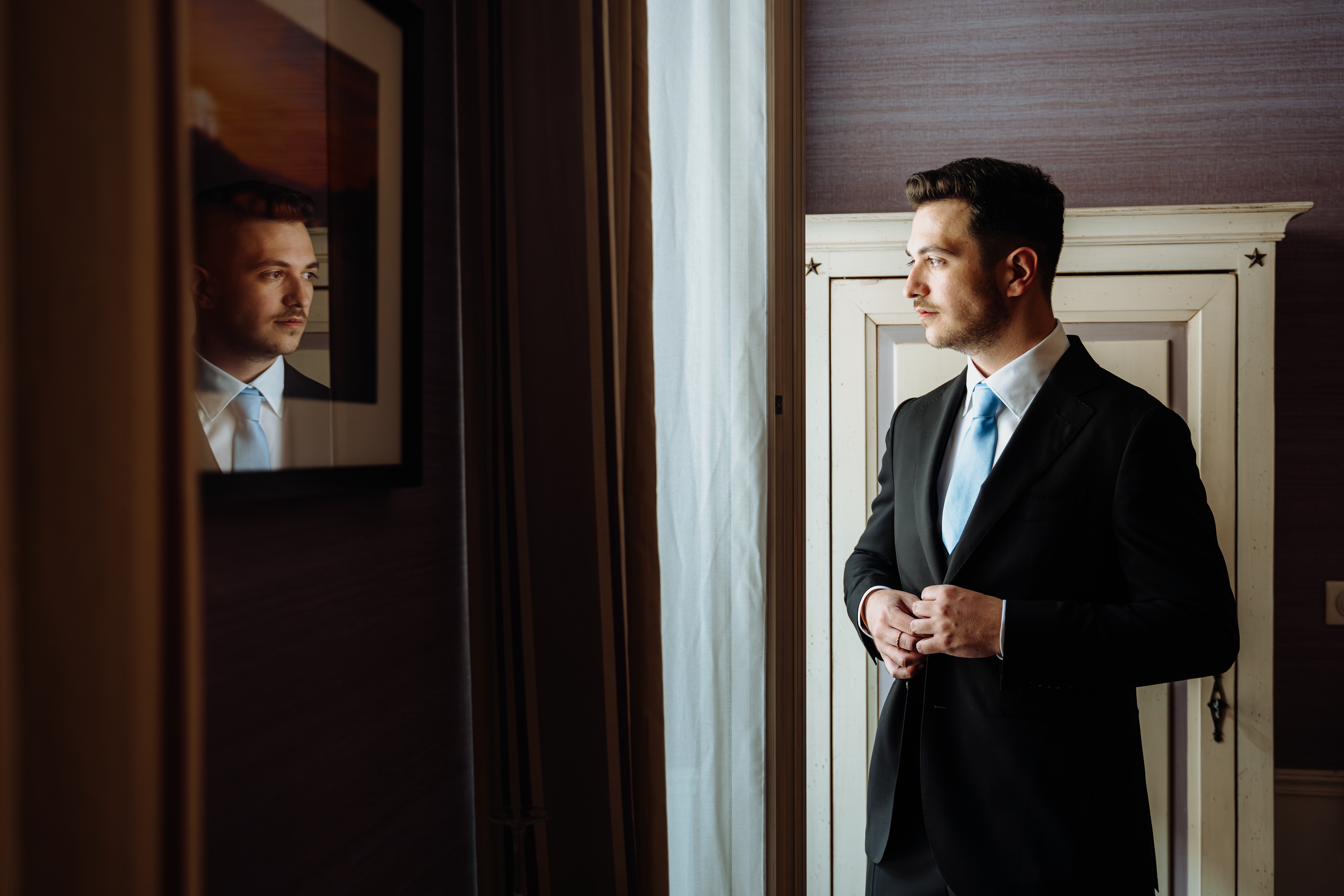 Photo du marié prise par Weddevent lors des préparatifs de son mariage, montrant le marié boutonnant sa veste et regardant par la fenêtre en apercevant son reflet dans le verre d'un cadre accroché au mur.